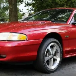 Czerwony Ford Mustang z lat 90. na podjeździe w Opolu, widok z przodu, z lekkim skrętem w lewo. Widoczne detale: felgi aluminiowe i logo na masce.