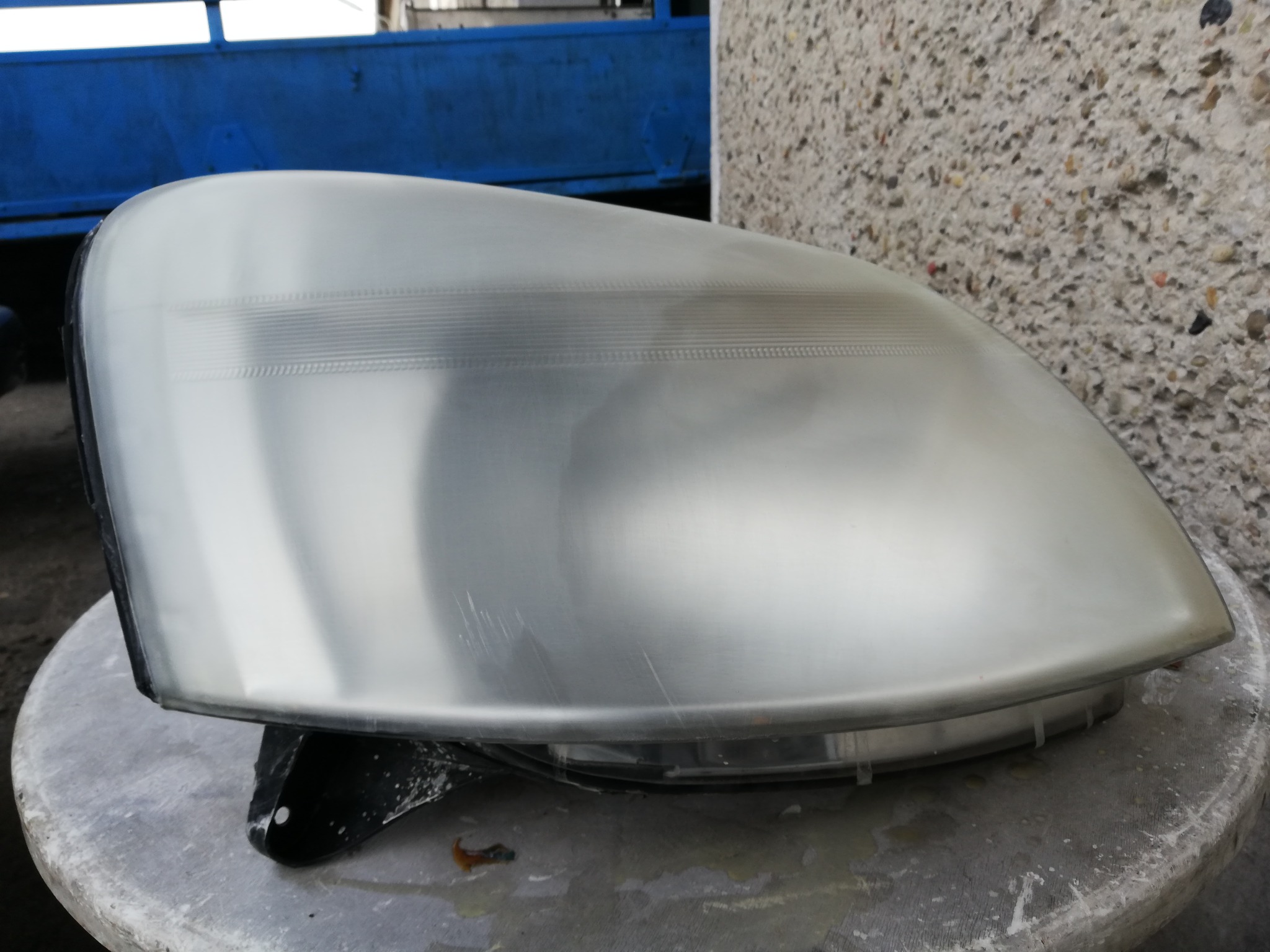 Reflektor samochodowy po polerowaniu, widoczne drobne rysy i ślady po paście polerskiej na powierzchni, w tle betonowy mur i fragment niebieskiej konstrukcji.