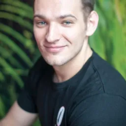 Portret uśmiechniętego mężczyzny o niebieskich oczach w czarnej koszulce z małym logo na tle zielonej ściany z motywem roślinnym.