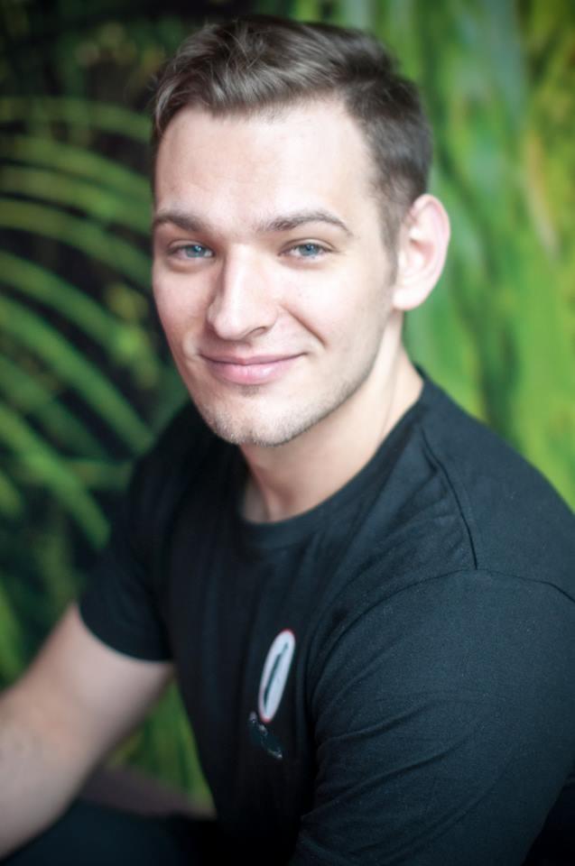 Portret uśmiechniętego mężczyzny o niebieskich oczach w czarnej koszulce z małym logo na tle zielonej ściany z motywem roślinnym.