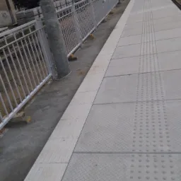 Nowy peron kolejowy z szarych płyt chodnikowych o fakturze rombów i wypukłych punktów, zabezpieczony białą metalową barierką i betonowym słupem oświetleniowym.