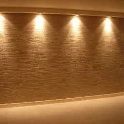 Beżowa ściana z fakturą i oświetleniem punktowym, widoczne gniazdka elektryczne, wykończenie podłogi w jasnym odcieniu.