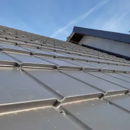 Szare, metalowe pokrycie dachowe z widocznymi mocowaniami, fragment konstrukcji dachu i błękitne niebo w tle.