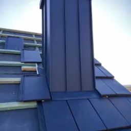 Montaż ciemnych dachówek ceramicznych na dachu z kominem w kolorze antracytowym, widoczne elementy konstrukcji drewnianej.