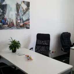 Nowoczesne biuro z białymi biurkami, czarnymi krzesłami i obrazem Times Square na ścianie, roślina doniczkowa na stole konferencyjnym.