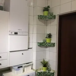 Biała łazienka z narożnymi półkami wykończonymi zieloną mozaiką, na których stoją doniczki z roślinami i szklanki, z pralką i piecem gazowym w tle.