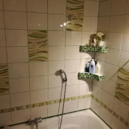 Białe płytki w łazience z dekoracyjnymi wstawkami w odcieniach zieleni, mozaikowe półki narożne z kosmetykami i kwiatami, widoczna bateria wannowa i słuchawka prysznicowa.