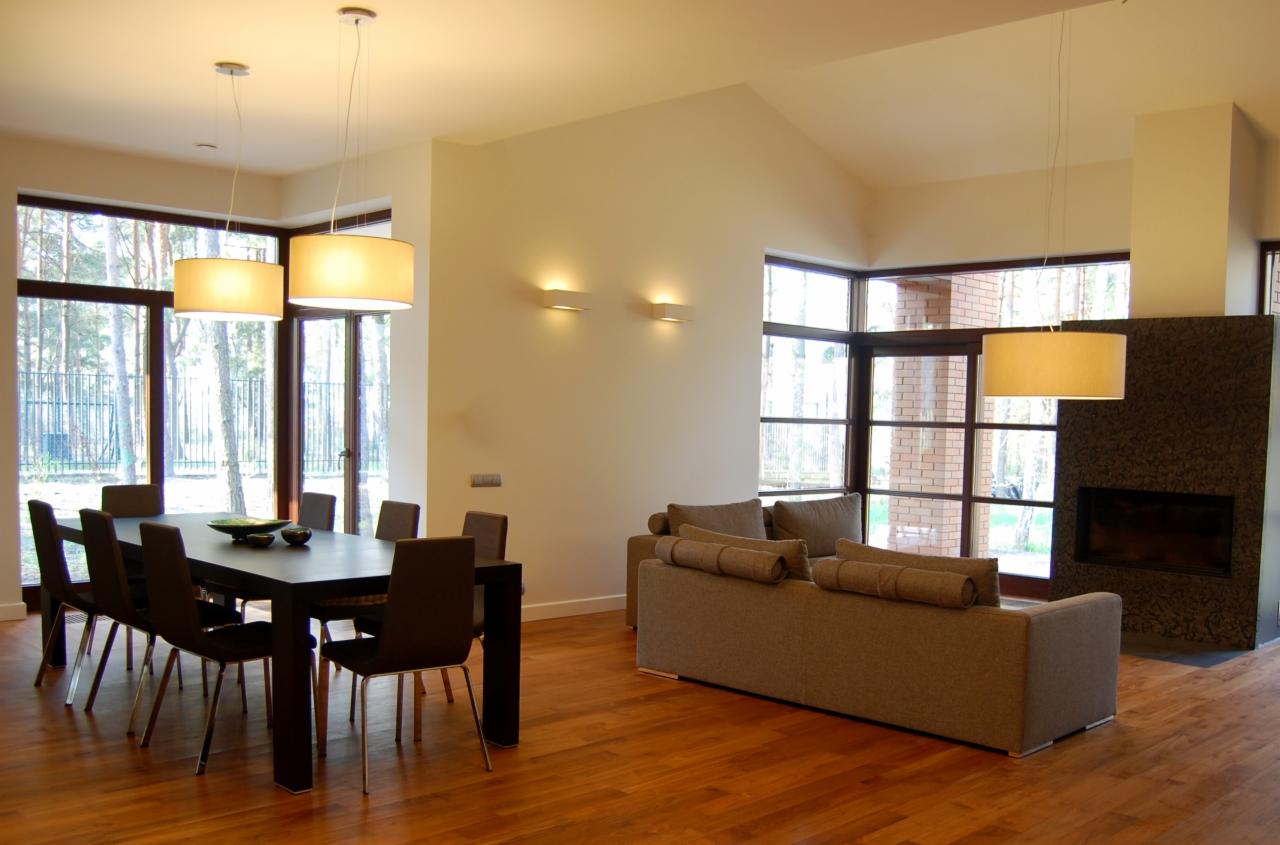 Jasne, przestronne wnętrze z drewnianą podłogą, dużym oknem z widokiem na las, stołem jadalnym z ciemnymi krzesłami, szarą sofą i kominkiem z ciemnej mozaiki.