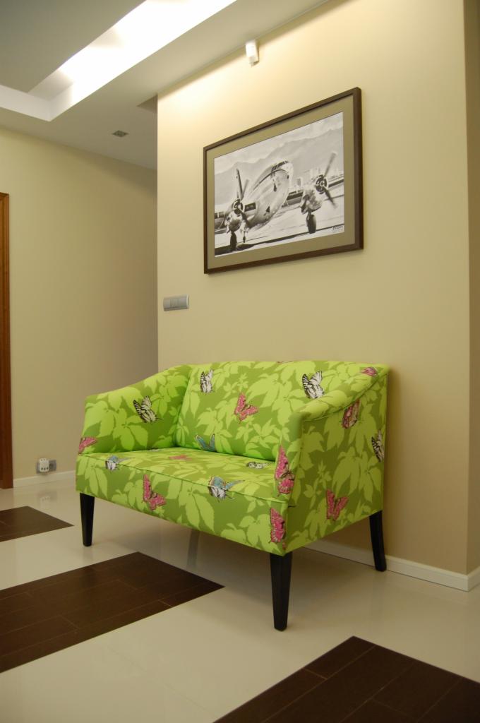 Jasnozielona sofa z motywem liści i motyli na tle beżowej ściany z czarno-białym zdjęciem samolotu w brązowej ramie, na podłodze płytki w szachownicę.