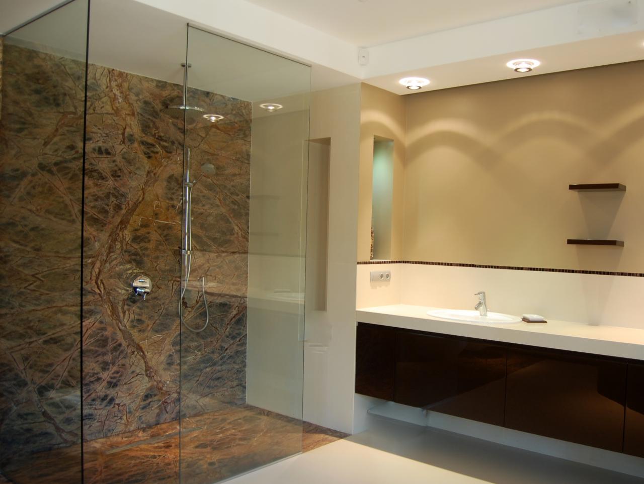 Nowoczesna łazienka z kabiną prysznicową typu walk-in, wykończoną marmurową okładziną z wyraźnym, naturalnym wzorem żyłkowania oraz długim blatem z ciemnobrązowymi szafkami podwieszanymi.