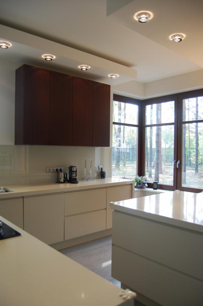 Nowoczesna kuchnia z beżowymi szafkami, ciemnobrązowymi szafkami wiszącymi i wyspą na pierwszym planie, widok na las przez duże okno.