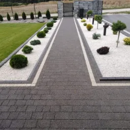 Ułożona szara kostka brukowa tworząca ścieżkę przez ogród z białym żwirem, ozdobnymi krzewami i lampami ogrodowymi, z trawnikiem po lewej stronie i ogrodzeniem w tle.