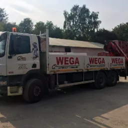 Ciężarówka firmy WEGA z materiałami budowlanymi na pace, zaparkowana na placu, widoczna grafika robota na kabinie, w tle drzewa i magazyn.