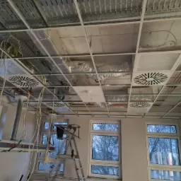Widok na sufit podwieszany w trakcie montażu instalacji wentylacyjnej z widocznymi kanałami wentylacyjnymi, okrągłymi nawiewnikami, przewodami elektrycznymi, drabina monterska i fragmentem...