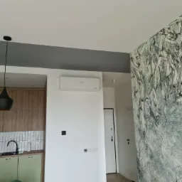 Wnętrze mieszkania z zamontowanym klimatyzatorem Mitsubishi na białej ścianie, widoczna kuchnia z zielonymi szafkami i wzorzysta tapeta w odcieniach błękitu i szarości.