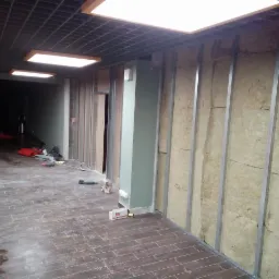 Wnętrze w trakcie prac: ściana z wełną mineralną między metalowymi profilami, podłoga wyłożona płytkami imitującymi drewno, widoczne narzędzia i materiały budowlane, sufit podwieszany z rastrem...
