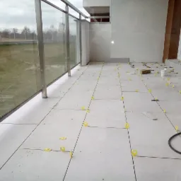 Balkon w trakcie układania jasnych, kwadratowych płytek z użyciem ż&oacute;łtych krzyżyk&oacute;w dystansowych, widoczne narzędzia glazurnicze i fragment krajobrazu za szklaną balustradą.