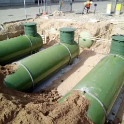 Montaż szamba ekologicznego: trzy zielone zbiorniki cylindryczne z zielonymi włazami w wykopie, częściowo zasypane piaskiem, w tle budynek i pracownik w kamizelce kopiący ziemię.