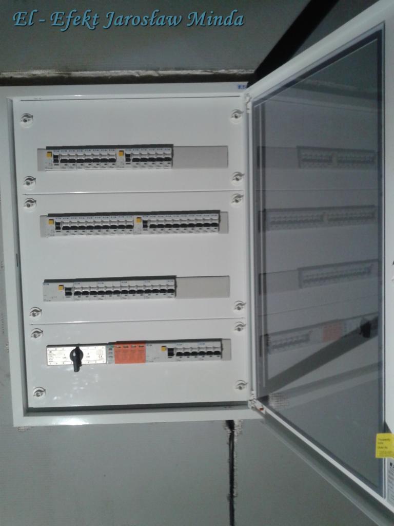 Otwarta skrzynka rozdzielcza z czterema rzędami zabezpieczeń elektrycznych, widoczny wyłącznik główny i zabezpieczenia różnicowoprądowe, zamontowana na szarej ścianie.