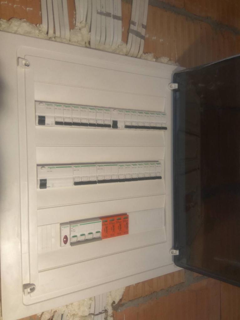 Otwarta skrzynka bezpiecznikowa z widocznymi zabezpieczeniami różnicowoprądowymi i nadprądowymi, zamontowana na surowej ścianie z cegły.