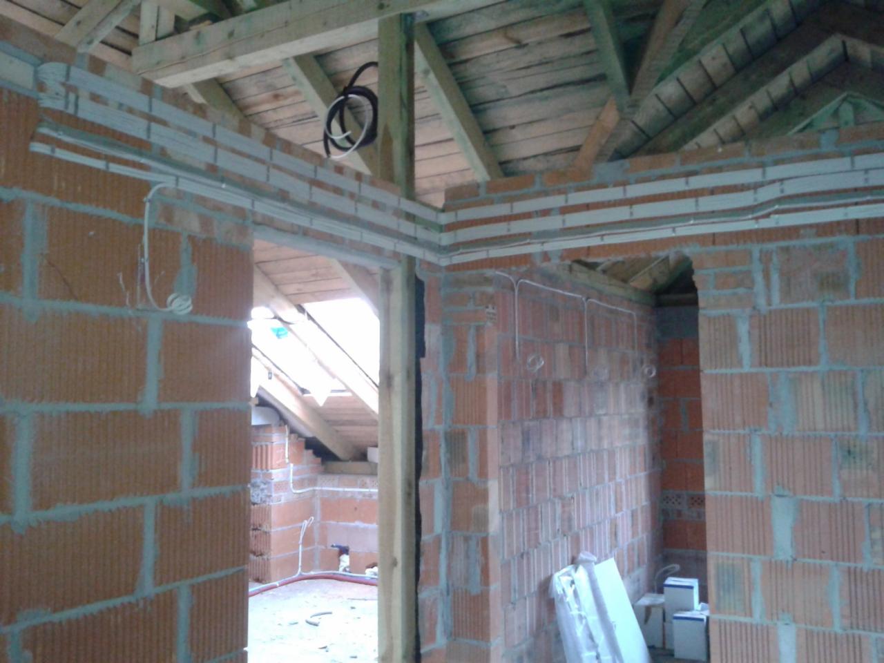 Wewnętrzne ściany z czerwonej cegły w surowym stanie, z widocznymi poziomymi listwami instalacyjnymi i przewodami elektrycznymi, drewniana konstrukcja dachu w tle, widok na poddasze przez otwór...