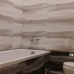 Wykończona łazienka z wanną obłożoną ciemnymi płytkami imitującymi drewno i jasnymi płytkami na ścianach z motywem fal, chromowana bateria umywalkowa, toaleta w tle.