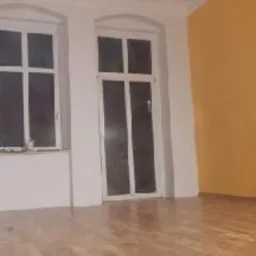 Pusty pokój po odświeżeniu: parkiet, żółta ściana, białe okna i sufit ze sztukaterią.