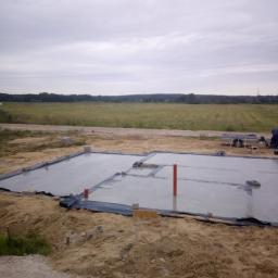 ARDOM - Świeżo wylany fundament pod budynek, pokryty folią ochronną, z widocznymi pionowymi rurami kanalizacyjnymi i fragmentem krajobrazu polnego w tle.