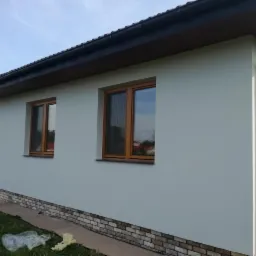 Świeżo wykończona elewacja domu jednorodzinnego z widocznymi drewnianymi oknami i fragmentem dachu z rynnami. Na dole elewacji widoczna okładzina imitująca kamień.