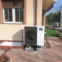 Biała pompa ciepła Mitsubishi Zubadan R32 zamontowana przy ścianie domu z brązowymi okiennicami, widoczna kostka brukowa i elementy ogrodu.