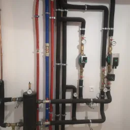 Złożona instalacja grzewcza z rurami w izolacji (czerwonej, niebieskiej i czarnej), manometrami i pompami obiegowymi na białej ścianie, widoczny fragment podłogi z jasnych płytek.