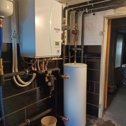 Eko - DOM - Biała pompa ciepła Ecodan z podłączonym zasobnikiem ciepłej wody, rurami i zaworami, zamontowana na ścianie wyłożonej czarnymi płytkami, widoczne także rury grzewcze i wiadro na podłodze.