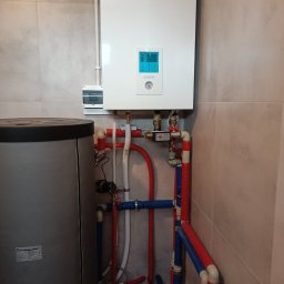 Eko - DOM - Biały wiszący piec gazowy Panasonic Aquarea z niebieskim wyświetlaczem, podłączony do instalacji hydraulicznej z rurami w kolorach czerwonym i niebieskim, obok szary zbiornik, na tle płytek...