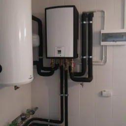 Instalacja kotła gazowego Vaillant z rurami w izolacji termicznej i zasobnikiem Galmet, zamontowana na ścianie wyłożonej białymi płytkami, widoczny licznik i zawory.