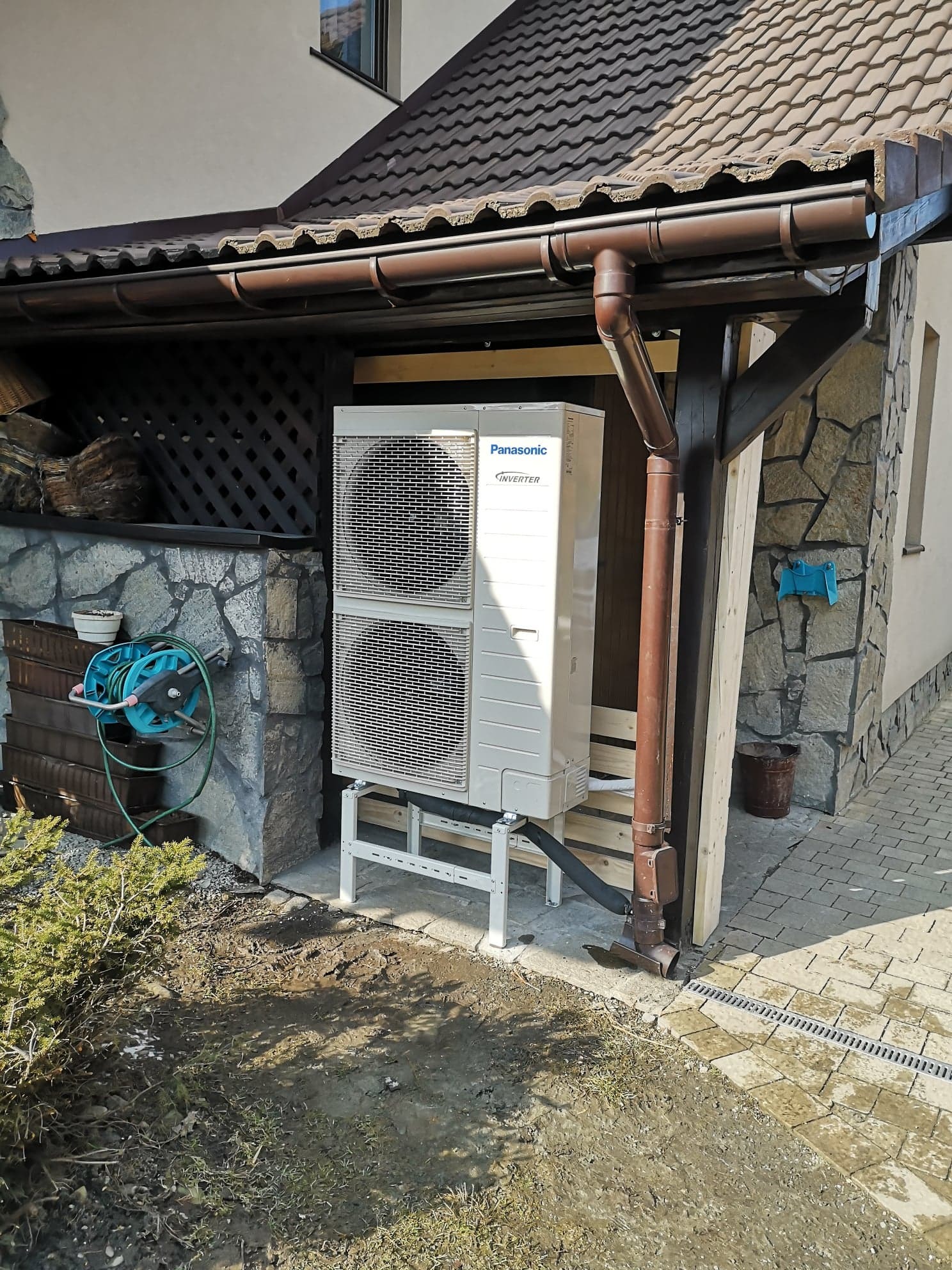 Zewnętrzna jednostka pompy ciepła Panasonic Inverter zamontowana na białym metalowym stelażu przy ścianie domu z kamienia łupanego i brązową rynną, widoczny fragment trawnika z młodymi roślinami.