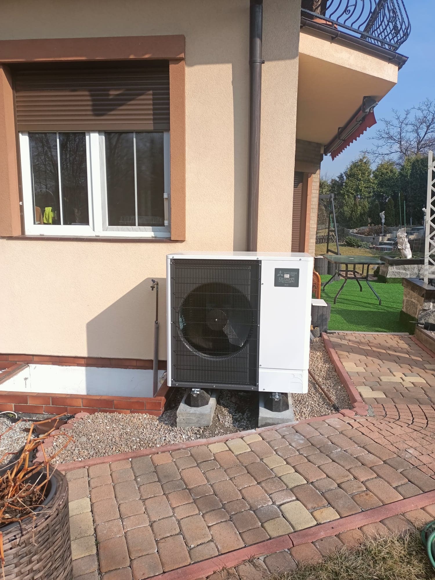 Biała pompa ciepła Mitsubishi Zubadan R32 zamontowana przy ścianie domu z brązowymi okiennicami, widoczna kostka brukowa i elementy ogrodu.
