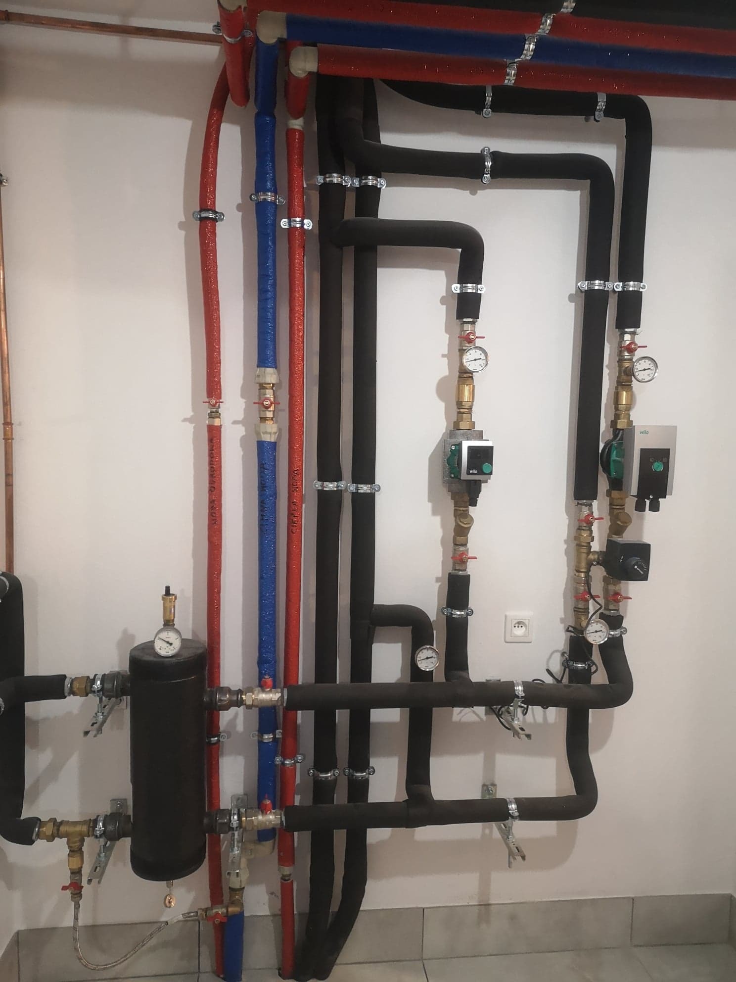 Złożona instalacja grzewcza z rurami w izolacji (czerwonej, niebieskiej i czarnej), manometrami i pompami obiegowymi na białej ścianie, widoczny fragment podłogi z jasnych płytek.