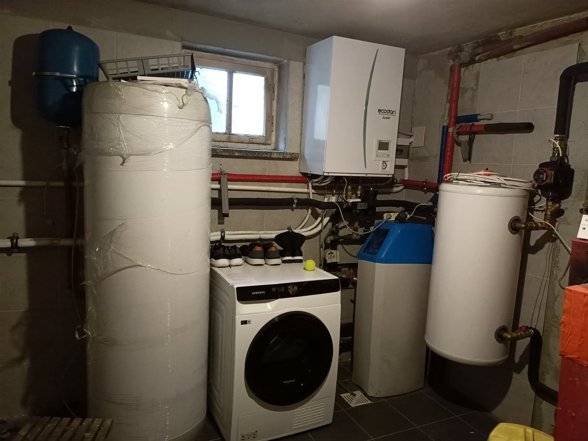 Piwnica z zainstalowanym kotłem gazowym Ecodan, zbiornikiem buforowym owiniętym folią, zmiękczaczem wody z niebieską pokrywą, suszarką do ubrań Samsung i niebieskim zbiornikiem ciśnieniowym.