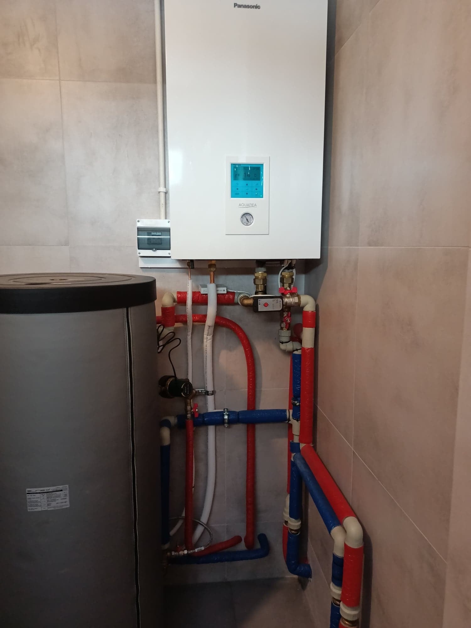 Biały wiszący piec gazowy Panasonic Aquarea z niebieskim wyświetlaczem, podłączony do instalacji hydraulicznej z rurami w kolorach czerwonym i niebieskim, obok szary zbiornik, na tle płytek...