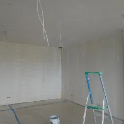 Pokój w trakcie przygotowania do malowania; ściany po nałożeniu gładzi, zabezpieczona podłoga folią, widoczna drabina aluminiowa i wystające przewody elektryczne z sufitu.
