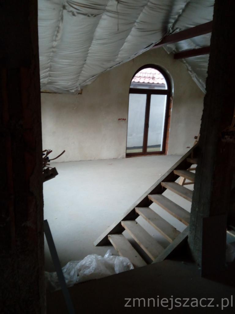 Poddasze w trakcie adaptacji, widoczne ocieplenie dachu, surowa ściana z oknem łukowym i drewniane schody prowadzące na wyższy poziom.