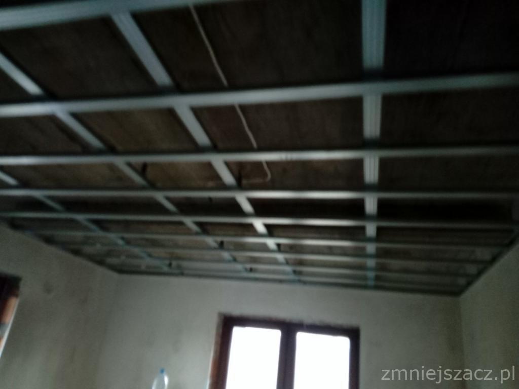 Sufit podwieszany w trakcie montażu z widoczną konstrukcją metalową i częściowo zamontowanymi płytami, surowy stan wykończenia wnętrza.
