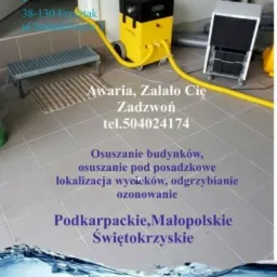 Żółto-czarny osuszacz przemysłowy na kółkach z podłączonymi żółtymi wężami, obok srebrny grzejnik na kółkach, szara podłoga z płytek, w tle drzwi i schody z metalową kratką, na zdjęciu widoczny...