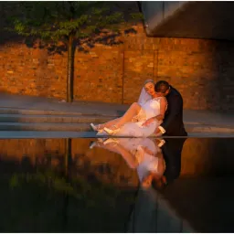 Panna młoda w białej sukni i welonie siedzi z panem młodym w granatowym garniturze na tle ceglanej ściany, ich odbicie widoczne w wodzie.