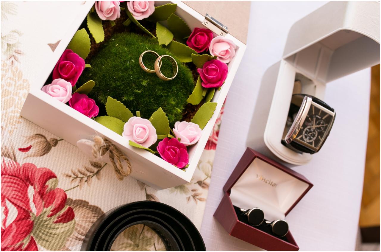 Obrączki ślubne w białym pudełku wypełnionym mchem i otoczone różowymi i czerwonymi piankowymi różyczkami, obok zegarek w pudełku i spinki do mankietów.