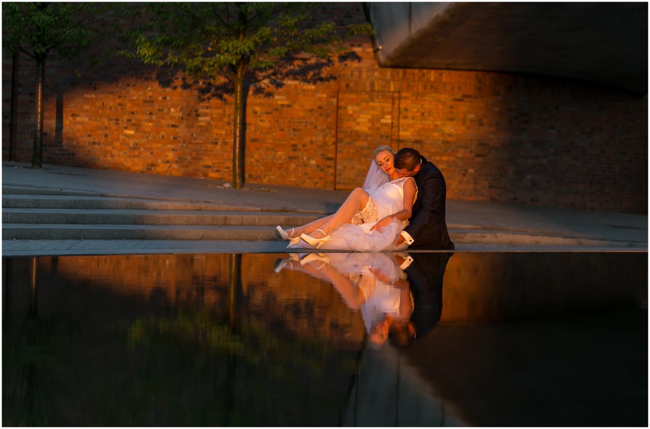 Panna młoda w białej sukni i welonie siedzi z panem młodym w granatowym garniturze na tle ceglanej ściany, ich odbicie widoczne w wodzie.