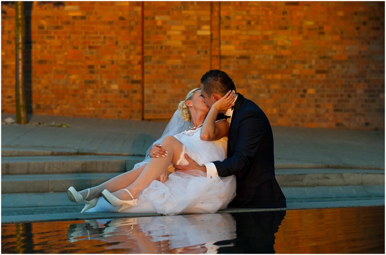 Panna młoda w krótkiej sukni ślubnej i białych szpilkach, siedząca na murku z odbiciem w wodzie, całowana przez pana młodego na tle ceglanej ściany.