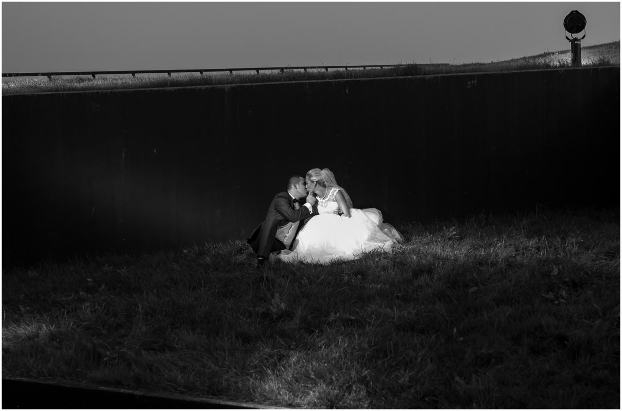 Czarno-białe zdjęcie pary młodej siedzącej na trawie i całującej się na tle ciemnego muru z betonowym ogrodzeniem i lampą w tle.