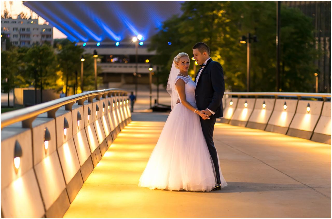 Para młoda trzymająca się za ręce na oświetlonym moście miejskim z nowoczesną architekturą w tle.