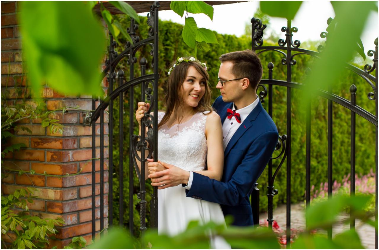 Para młoda w ogrodzie, panna młoda w białej sukni i wianku, pan młody w niebieskim garniturze i czerwonej muszce, uśmiechają się do siebie, trzymając się za ręce na tle metalowej bramy i ceglanego...